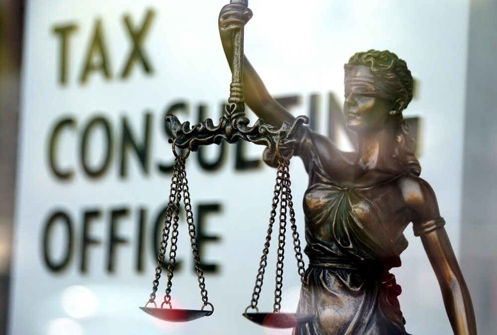 A imagem mostra uma estátua da deusa da justiça (conhecida como Justitia ou Lady Justice) em primeiro plano, com uma balança em uma das mãos e os olhos vendados. Ao fundo, desfocado, aparece o letreiro com os dizeres “TAX CONSULTING OFFICE”, que em português significa “ESCRITÓRIO DE CONSULTORIA TRIBUTÁRIA”.