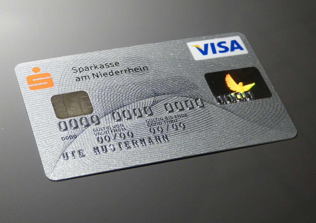 A imagem mostra um cartão de crédito prateado da bandeira Visa, emitido pelo banco Sparkasse am Niederrhein (uma instituição financeira da Alemanha). O cartão possui um chip na parte esquerda, usado para pagamentos com chip e senha. Há um holograma com a imagem de um pássaro dourado em voo, no lado direito. O nome impresso no cartão é UTE MUSTERMANN, que é um nome genérico usado como exemplo na Alemanha (equivalente a “Fulano de Tal” no Brasil). Os números do cartão e as datas de validade estão como placeholders (00/00 e 00/99), indicando que se trata de um modelo de demonstração, não um cartão real. O fundo possui um padrão de linhas curvas e texturas para dificultar falsificações e dar um aspecto sofisticado.
