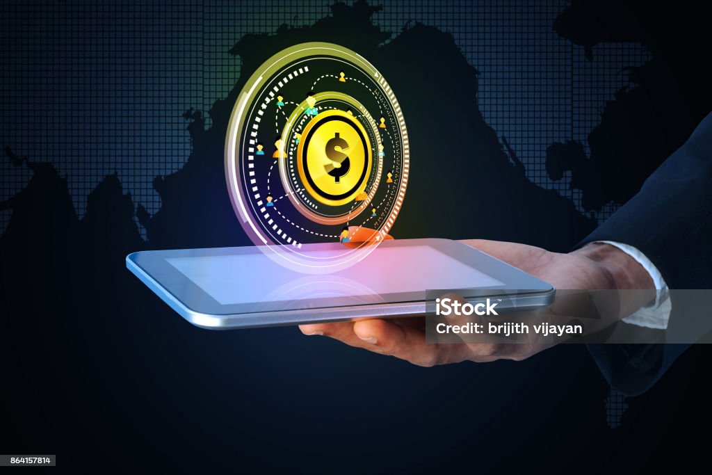 A imagem mostra uma mão segurando um tablet, de onde surge uma projeção holográfica futurista. No centro da projeção aparece um círculo dourado com o símbolo de um dólar ($), cercado por elementos digitais em formato circular, lembrando uma interface de tecnologia avançada. O fundo é escuro, com um mapa-múndi estilizado em grade digital, transmitindo a ideia de globalização, finanças digitais e inovação tecnológica. A cena remete a moedas digitais, criptomoedas, transações financeiras eletrônicas ou sistemas de pagamento digitais, representando a fusão entre tecnologia e economia moderna.
