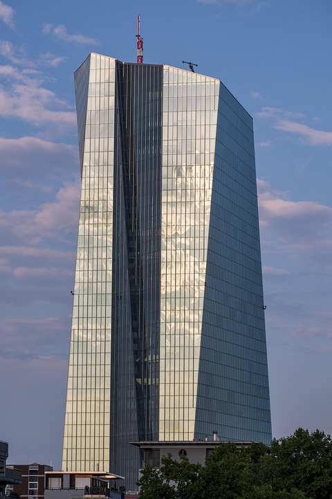 A imagem mostra um prédio moderno e alto, com fachada de vidro espelhado. A construção tem um design arquitetônico ousado, levemente inclinado e com linhas assimétricas, o que lhe dá um aspecto imponente e futurista. O reflexo das nuvens no vidro cria um efeito visual elegante, mesclando o edifício com o céu. No topo da torre, é possível ver uma antena vermelha e outros equipamentos. Ao redor da base aparecem algumas construções menores e árvores, sugerindo que o edifício está localizado em uma área urbana. Esse tipo de prédio transmite uma sensação de modernidade, poder econômico e inovação arquitetônica.