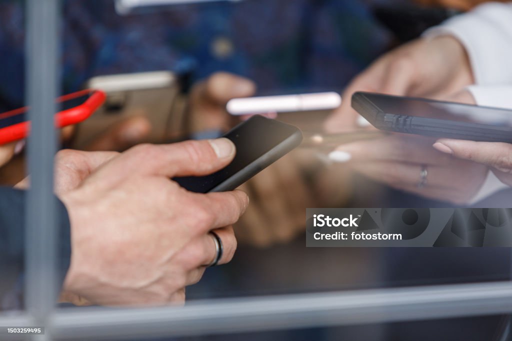 A imagem mostra um grupo de pessoas segurando e utilizando seus smartphones. As mãos estão em destaque, com diferentes modelos de celulares sendo manuseados, alguns com capinhas protetoras. O foco está nas interações digitais, transmitindo a ideia de conectividade, tecnologia e comunicação móvel. O ambiente parece ser coletivo, sugerindo uma situação onde várias pessoas estão concentradas em seus aparelhos ao mesmo tempo.