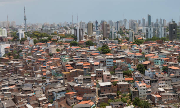 A imagem mostra uma cena urbana marcada pelo contraste social. Na parte inferior e central, aparecem diversas casas simples e construções irregulares, muito próximas umas das outras, típicas de áreas de periferia ou favela. Os telhados são, em sua maioria, de telhas vermelhas ou de materiais improvisados. Ao fundo, destaca-se uma paisagem com vários prédios altos e modernos, de padrão mais elevado, representando uma região mais desenvolvida da cidade. Esse contraste entre as moradias populares em primeiro plano e os edifícios altos no horizonte evidencia a desigualdade socioeconômica presente nos grandes centros urbanos do Brasil.