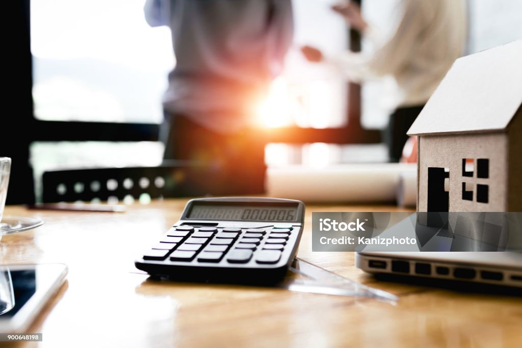 A imagem mostra uma mesa de trabalho com itens relacionados a finanças e planejamento imobiliário. Em primeiro plano, há uma calculadora, parte de um notebook e uma maquete em miniatura de uma casa de papelão, simbolizando imóveis ou financiamento habitacional. Também é possível ver um copo e um tablet parcialmente à esquerda. Ao fundo, desfocados, aparecem duas pessoas em pé conversando, reforçando a ideia de reunião ou negociação. A luz do sol entra pela janela, criando um ambiente iluminado e profissional. Essa cena transmite conceitos ligados a financiamento imobiliário, compra de imóveis, planejamento financeiro, investimentos e cálculos de viabilidade econômica.