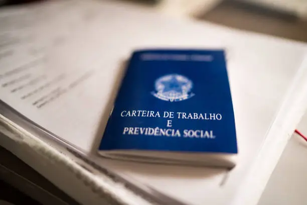 A imagem mostra uma Carteira de Trabalho e Previdência Social (CTPS), documento oficial brasileiro de capa azul. Ela está apoiada sobre algumas folhas de papel, provavelmente documentos, em uma mesa. A carteira aparece em destaque no centro da foto, levemente inclinada, e o foco está na capa onde se lê claramente o título em letras brancas. Esse documento é utilizado para registrar vínculos empregatícios, direitos trabalhistas e previdenciários no Brasil.