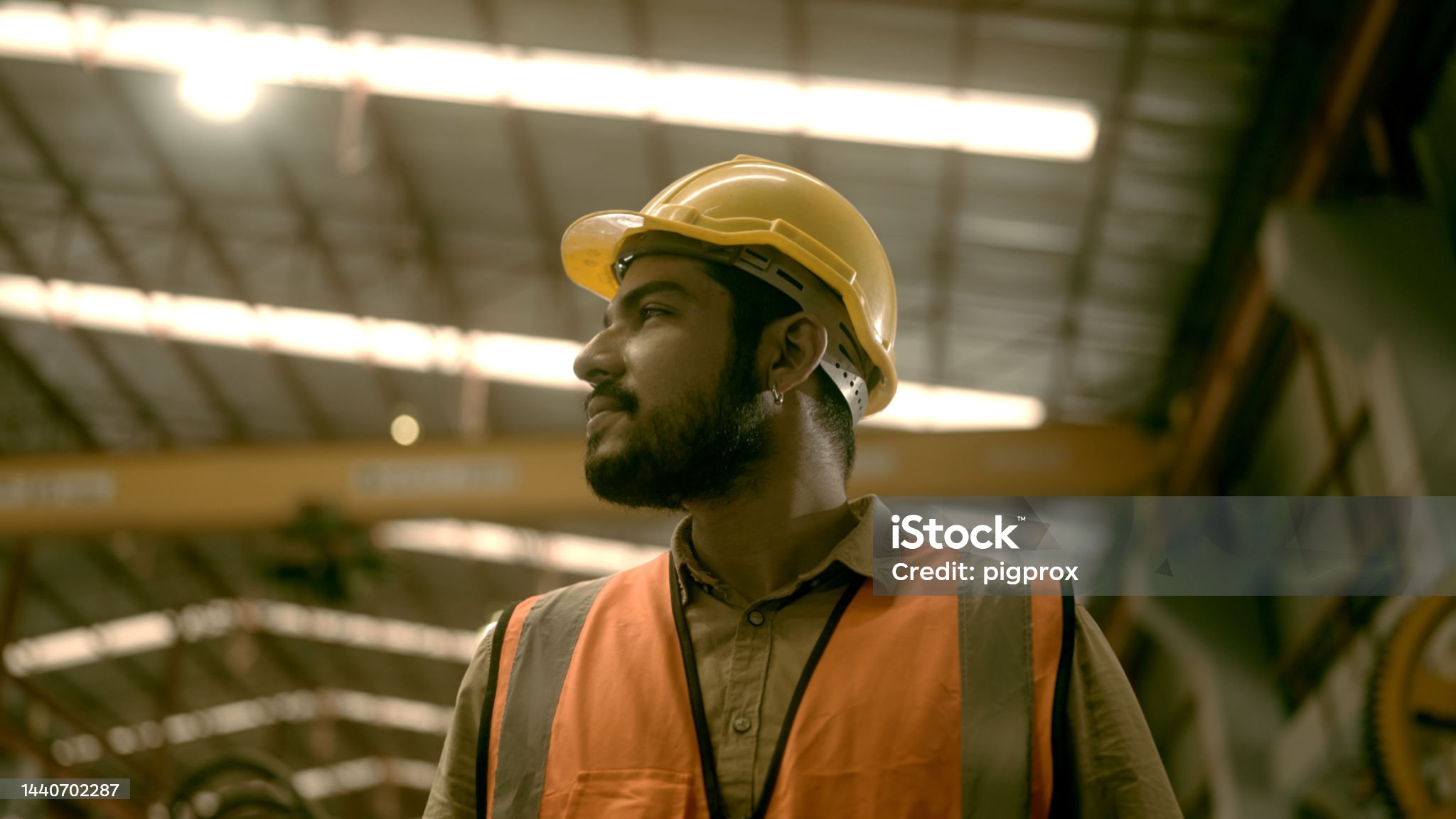 A imagem mostra um homem em um ambiente industrial ou de construção. Ele está usando um capacete de segurança amarelo e um colete de alta visibilidade laranja, que são equipamentos de proteção individual (EPIs) típicos para trabalhadores em obras ou fábricas. O homem tem barba e usa brinco, está olhando para o lado com uma expressão séria e concentrada. O fundo mostra uma estrutura metálica com iluminação artificial, sugerindo que ele está dentro de um galpão, armazém ou indústria.
