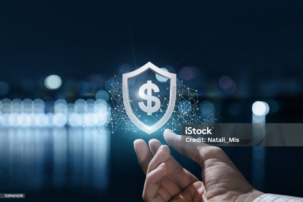 A imagem mostra uma mão estendida para cima, como se estivesse segurando ou oferecendo algo. Acima da mão, há um ícone digital em formato de escudo, no centro do qual aparece o símbolo de cifrão ($). O escudo é cercado por linhas e pontos luminosos interconectados, lembrando uma rede digital ou tecnológica. O fundo está desfocado, com luzes borradas que sugerem um cenário noturno urbano, possivelmente próximo à água. Essa composição transmite a ideia de segurança financeira, proteção de dinheiro, investimentos seguros, tecnologia aplicada às finanças (fintechs, blockchain, cibersegurança) ou ainda planejamento e proteção patrimonial.