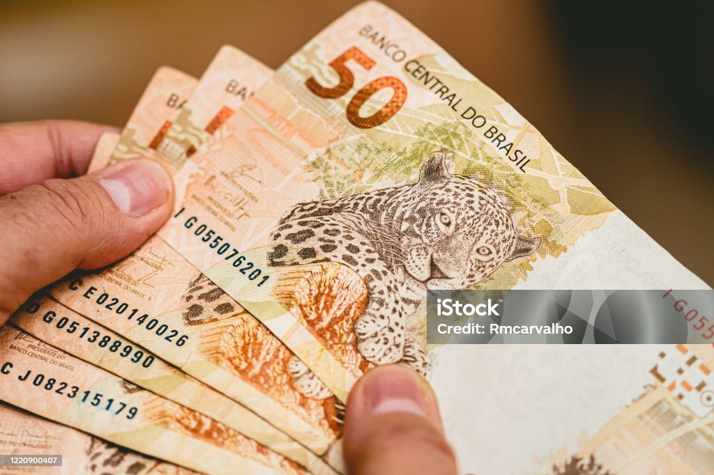 A imagem mostra uma mão segurando várias cédulas de 50 reais do Brasil. As notas estão em leque, destacando a parte frontal onde aparece a ilustração de uma onça-pintada, animal símbolo da cédula. Na parte superior, vê-se a inscrição “Banco Central do Brasil” e o número 50 em destaque, indicando o valor da nota. As cédulas têm tons predominantemente alaranjados e marrons, com detalhes de segurança visíveis, como números de série impressos. A cena transmite a ideia de dinheiro em espécie, finanças ou transações monetárias.