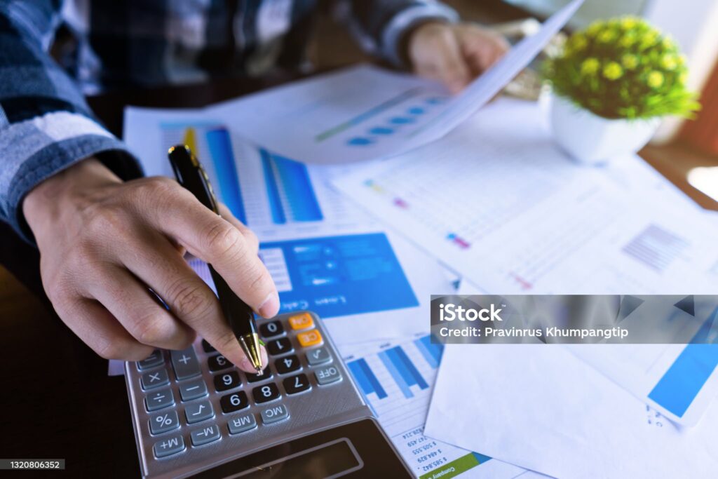A imagem mostra uma pessoa utilizando uma calculadora enquanto segura uma caneta, em meio a diversos documentos financeiros e gráficos impressos. O foco principal está na mão que digita na calculadora, transmitindo a ideia de controle de gastos, planejamento ou análise de finanças. Sobre a mesa, estão espalhados relatórios com gráficos de barras e tabelas em tons de azul, sugerindo planilhas de orçamento, investimentos ou relatórios de desempenho. Ao fundo, um vaso com planta verde dá um contraste visual, equilibrando o ambiente de trabalho. Essa imagem representa bem gestão financeira, planejamento de orçamento, controle de dívidas ou tomada de decisão financeira.