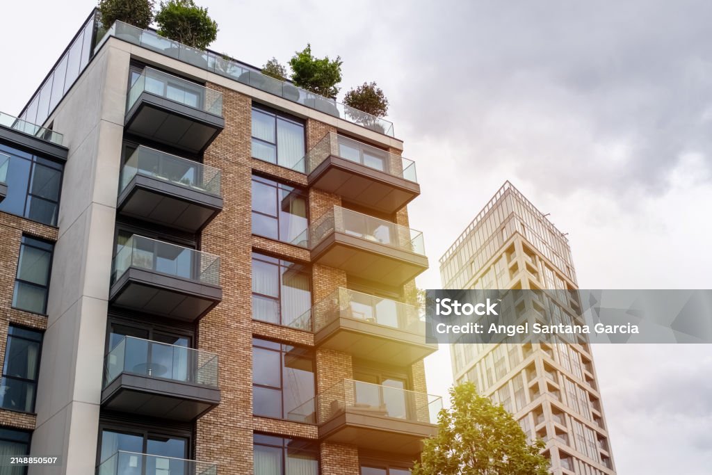A imagem mostra dois edifícios modernos, com design contemporâneo. O prédio em primeiro plano possui fachada de tijolos aparentes combinados com grandes janelas de vidro e varandas com guarda-corpo transparente. No topo do edifício, há um terraço com árvores e vegetação, dando um toque verde e sustentável à construção. Ao fundo, vê-se outro edifício mais alto e estreito, com fachada envidraçada e linhas verticais marcantes. O céu está nublado, com nuvens cinzentas, e a luz do sol incide suavemente sobre os prédios, criando um efeito de brilho e contraste na imagem.