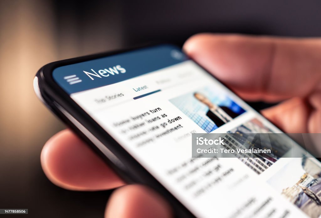 A imagem mostra uma pessoa segurando um smartphone que exibe uma página de notícias. Na tela do celular, na parte superior, aparece o título “News” em branco sobre um fundo azul. Logo abaixo, há abas de navegação, sendo possível identificar a aba “Latest” (Últimas) destacada. A tela exibe manchetes de artigos, acompanhadas por pequenas imagens ilustrativas. Embora o texto não esteja totalmente legível, percebe-se que são notícias atuais. A mão da pessoa está tocando ou rolando a tela, indicando que ela está navegando pelas informações. A foto foca no celular e na ação de leitura de notícias digitais, transmitindo a ideia de acesso rápido à informação e consumo de conteúdo online.